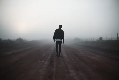 A person wearing dark clothing walks alone on a misty, dirt road near Khiron Clinics, with a fence running parallel on the right side and bushes on the left. The background is engulfed in thick fog, creating a moody and mysterious atmosphere.