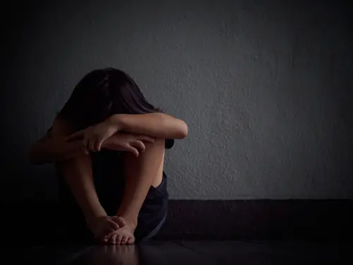 A person with long hair is sitting on the floor, curled up with their arms resting on their knees and head hidden. The scene is dimly lit, creating a somber and reflective mood against a plain, dark background, reminiscent of the introspective moments often experienced in trauma treatment at Khiron Clinics.