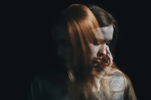 A woman with long hair appears in a double exposure effect. The two overlapping images show her looking down with a contemplative expression, one with her hand touching her face and the other by her side. Set against a dark background, this evocative image captures the introspective journey often facilitated at Khiron Clinics.