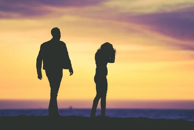 Silhouetted against a vibrant sunset, a man and woman stand on a beach. The man, on the left, appears to be walking towards the woman, who is on the right with her head bowed and hands covering her face, suggesting a contemplative or emotional moment indicative of someone seeking mental health treatment.