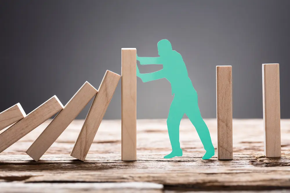 A teal silhouette of a person is shown preventing a series of wooden dominoes from falling. The individual leans against one upright domino, symbolizing the intervention and trauma treatment at a mental health facility to stop a chain reaction.