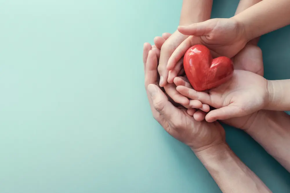 An adult hand and a child's hand are gently cupped together, holding a red heart-shaped object against a light blue background. This symbolizes care, love, and support across generations, reflecting the nurturing environment of trauma clinics like Khiron Clinics.