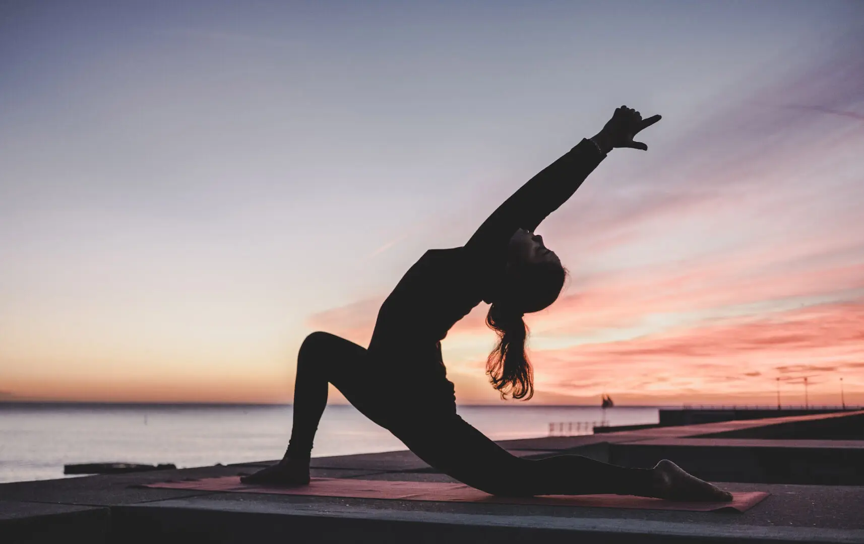 A person performs a yoga pose on a mat at the edge of a body of water during sunset, possibly as part of their mental health treatment. The sky is a gradient of soft pastel colors, with hints of orange, pink, and purple. The person stretches one leg forward and extends their arms upward, arching their back.