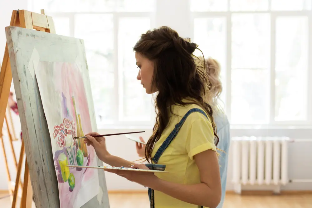 A woman with long brown hair is painting on a canvas in a well-lit studio, likely part of Khiron Clinics. She is wearing a yellow shirt and appears focused on her work. An easel holds her partially completed painting, and another person is faintly visible in the background.