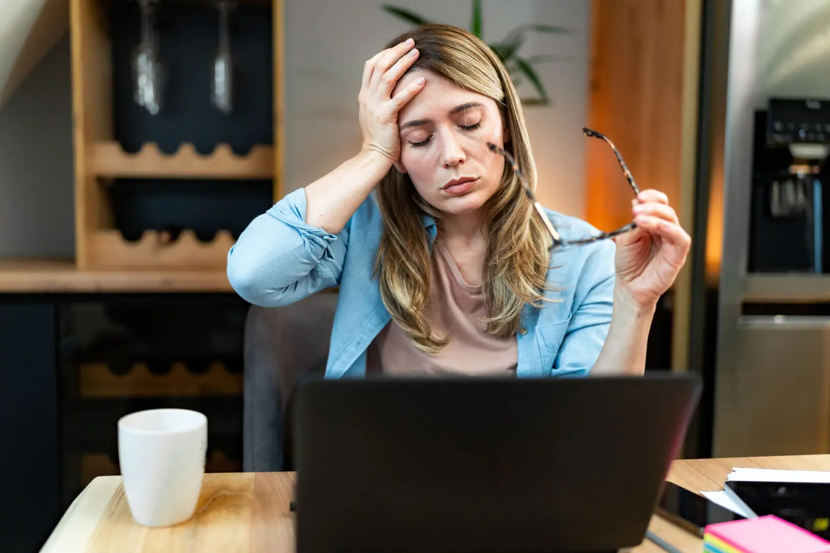 A woman sits at a table with a laptop in front of her, holding her forehead with one hand and glasses in the other. She looks tired or stressed. A white coffee mug is on the table. The background includes a wine rack and kitchen appliances, highlighting the pressures possibly eased by Khiron Clinics' trauma treatment.