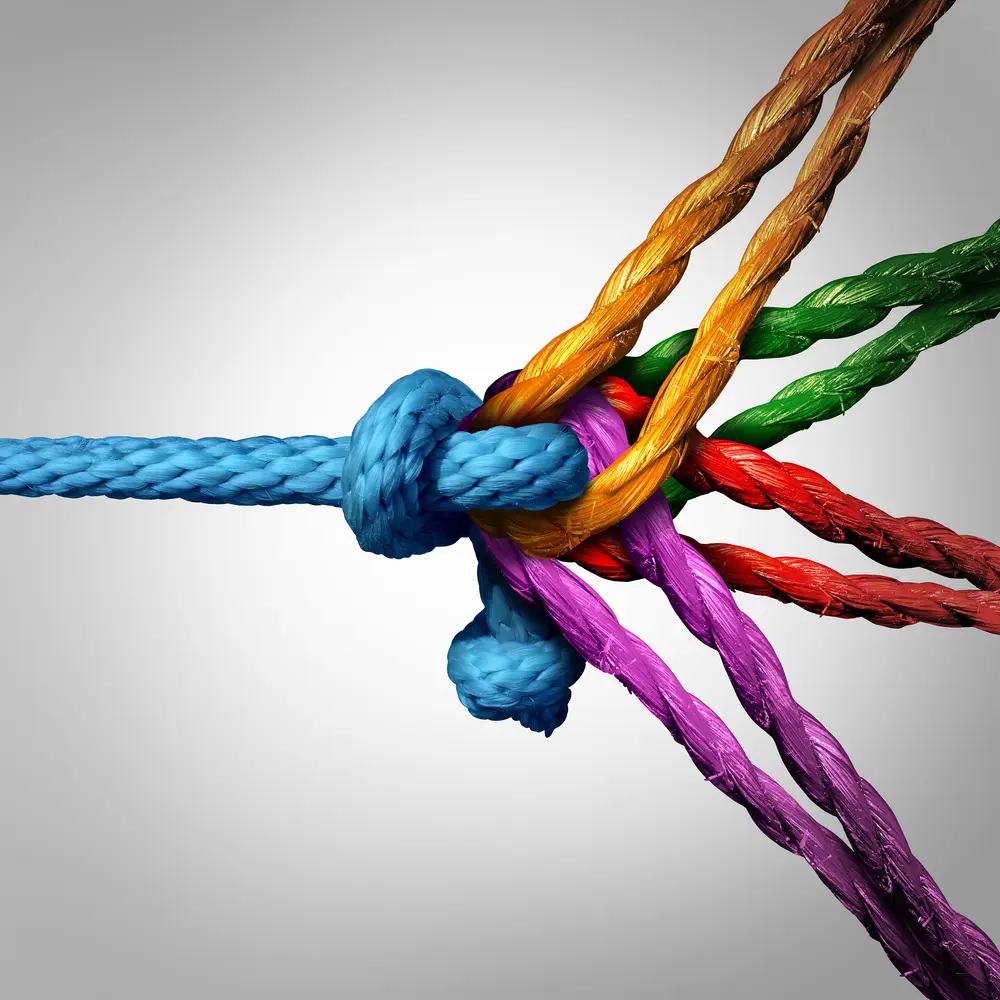 A close-up of multicolored ropes—blue, yellow, orange, green, red, and purple—tied together in a knot against a gradient gray background, symbolizing unity and cooperation within mental health treatment.