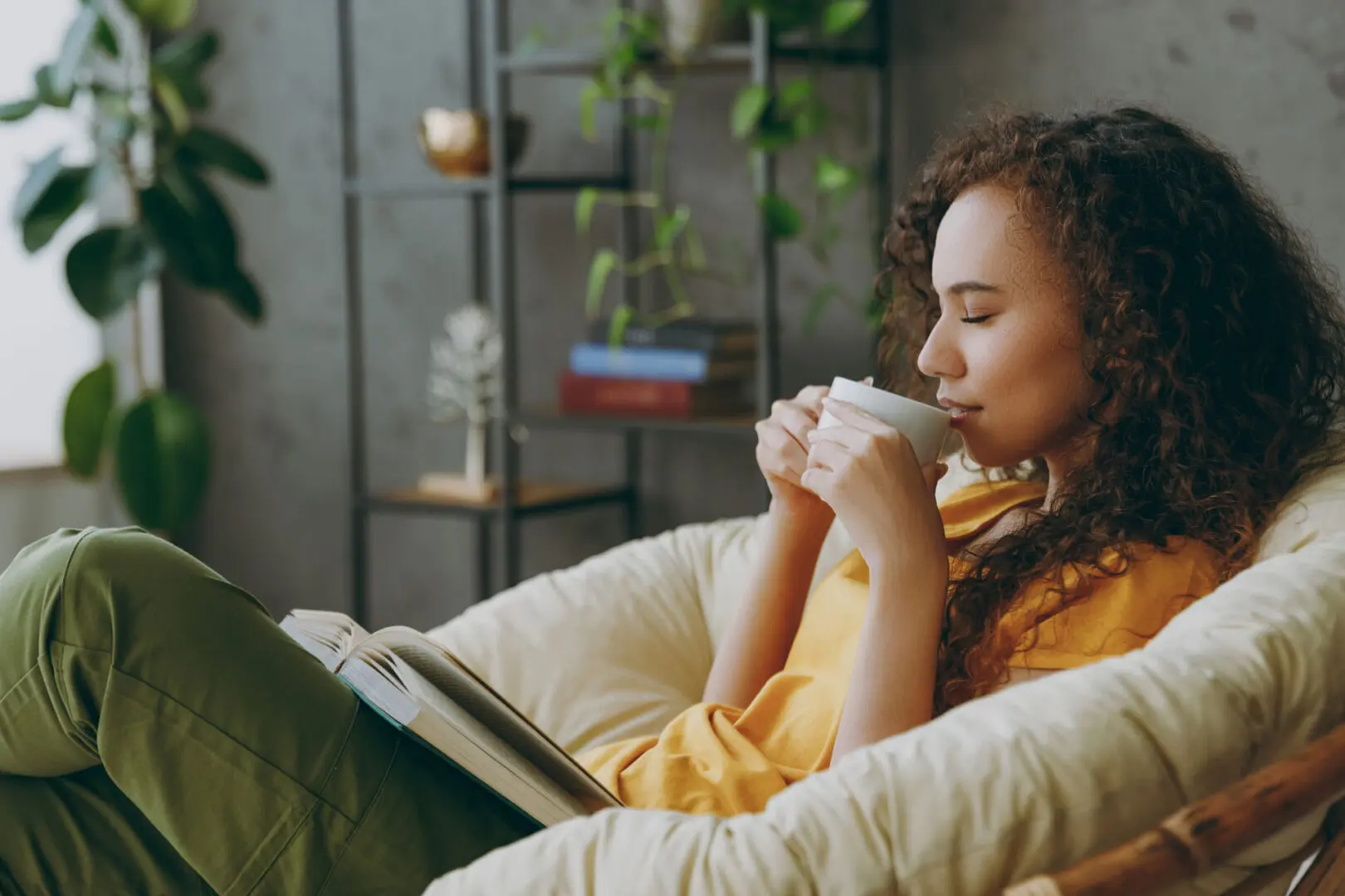 A person with curly hair sits comfortably in a cushioned chair, wearing a yellow top and green pants. They hold a cup close to their face, eyes closed, seemingly enjoying the aroma. An open book rests on their lap. The serene setting feels like a haven from the rigors of PTSD treatment at a trauma clinic.