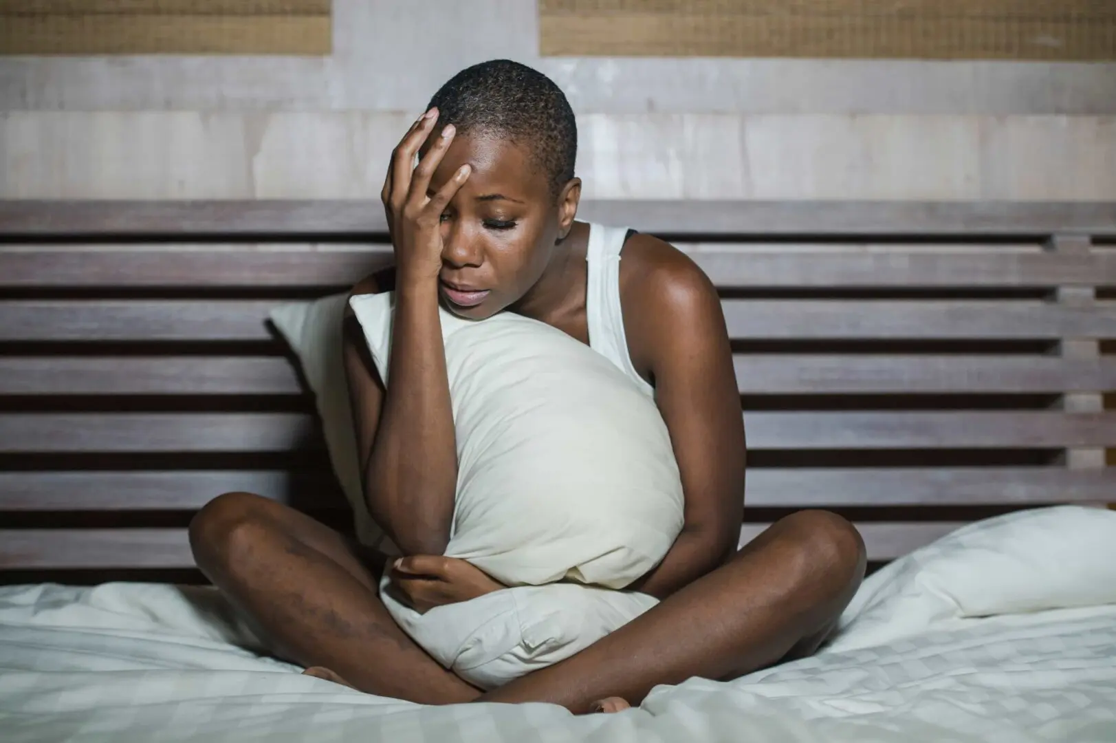 A person with short hair sits on a bed, clutching a pillow with one arm while resting their head in their other hand. They appear distressed or contemplative, perhaps reflecting on trauma treatment at Khiron Clinics, with a wooden headboard visible in the background.