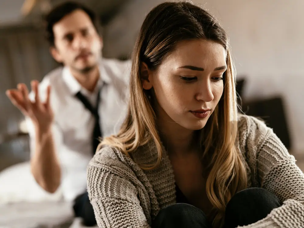 A woman with long hair sits in the foreground, looking down and appearing upset. A man in the background, wearing a shirt and tie, has his hand raised and seems frustrated. The image suggests a tense or emotional conversation, possibly set in a trauma clinic like Khiron Clinics.