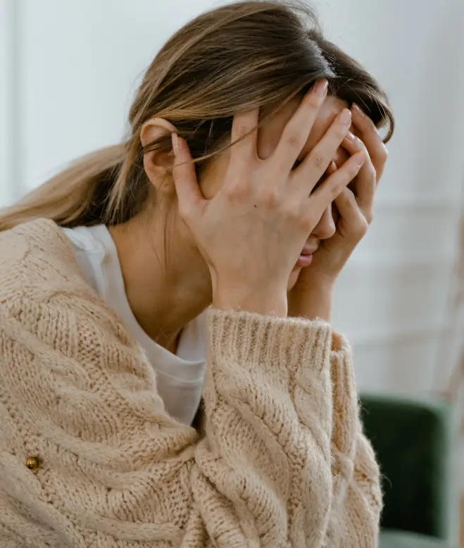 A person with light brown hair pulled back and wearing a beige knit sweater is sitting and holding their head in their hands, conveying a sense of stress or distress. Their face is partially obscured by their hands, suggesting they might benefit from mental health treatment at Khiron Clinics.