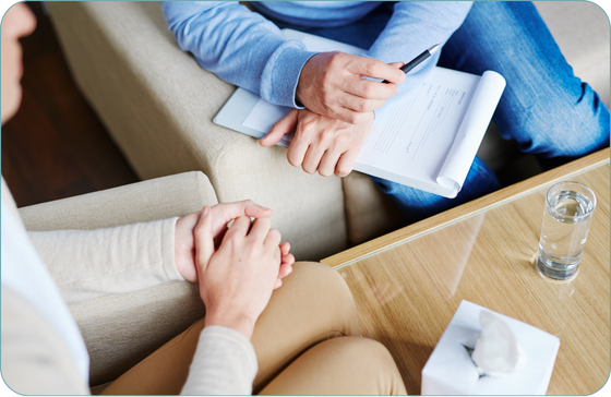 Two people are in a close conversation. One person is holding a pen and clipboard with papers, seated across from the other who has hands clasped in their lap. A table between them holds a glass of water and a tissue box. The setting suggests a mental health treatment environment, possibly for trauma treatment.