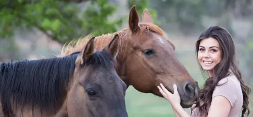 A woman with long dark hair smiles while petting the heads of two horses, one light brown and one dark brown, in an outdoor setting with green foliage in the background. This serene moment is part of her trauma treatment at a specialized clinic - using equine therapy