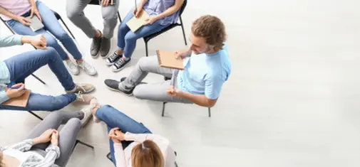 A group of people sits in a circle on chairs, some holding notebooks and pens. They are casually dressed, suggesting an informal meeting or group discussion at Khiron Clinics. The view is from above, focusing on their legs and not showing faces clearly, perhaps during a session for trauma treatment.