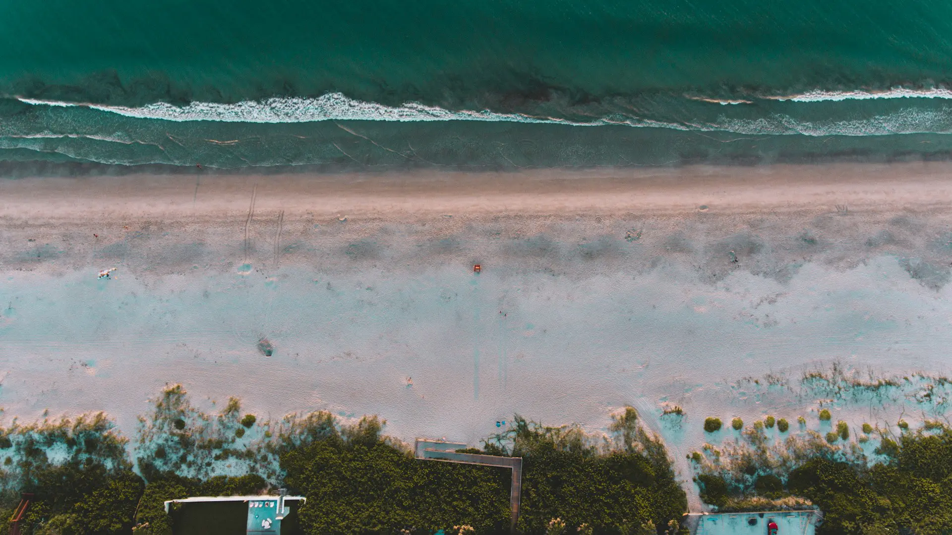 Aerial view of a beach with turquoise waves gently crashing on the sandy shore. Green, grassy areas border the sand, and a few small structures are visible near the edge of the beach. A lone figure in a red outfit stands in the middle of the sand, perhaps contemplating what is trauma amidst nature's tranquility.