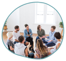 A group of people sits in a circle in a well-lit room with wooden floors and potted plants. They appear to be engaged in a mental health treatment session or group therapy. Some individuals have their arms crossed, while others use hand gestures as they speak.