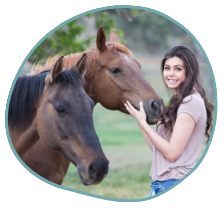 A woman with long dark hair is standing outdoors, smiling, and gently touching the face of a brown horse. Another brown horse is standing close by. The background, green with trees and grass, offers a serene setting that could aid in PTSD treatment - including equine therapy