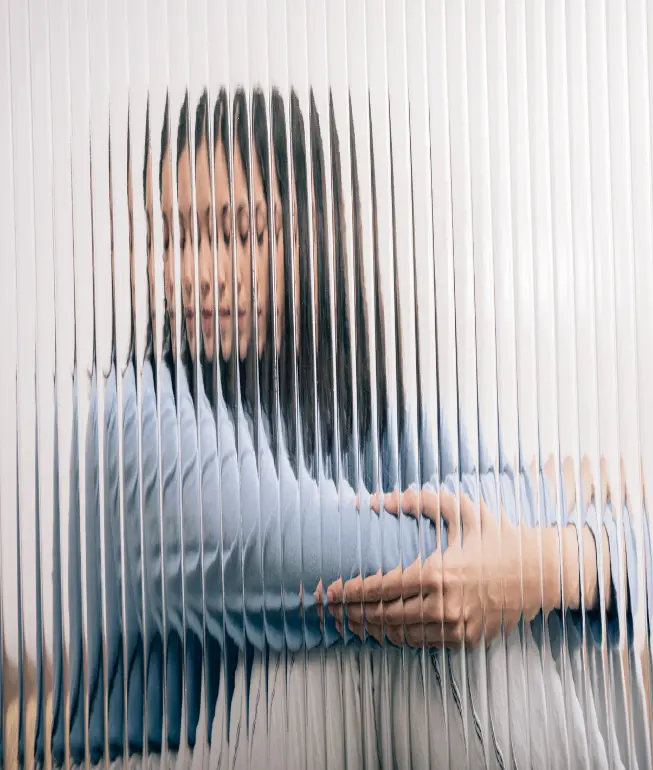 A person with long dark hair appears multiple times due to the distortion of a ribbed glass barrier. They are wearing a light blue sweater and have their arms crossed. The overall effect creates an abstract, surreal image, reminiscent of the fragmented perceptions treated at Khiron Clinics for PTSD.