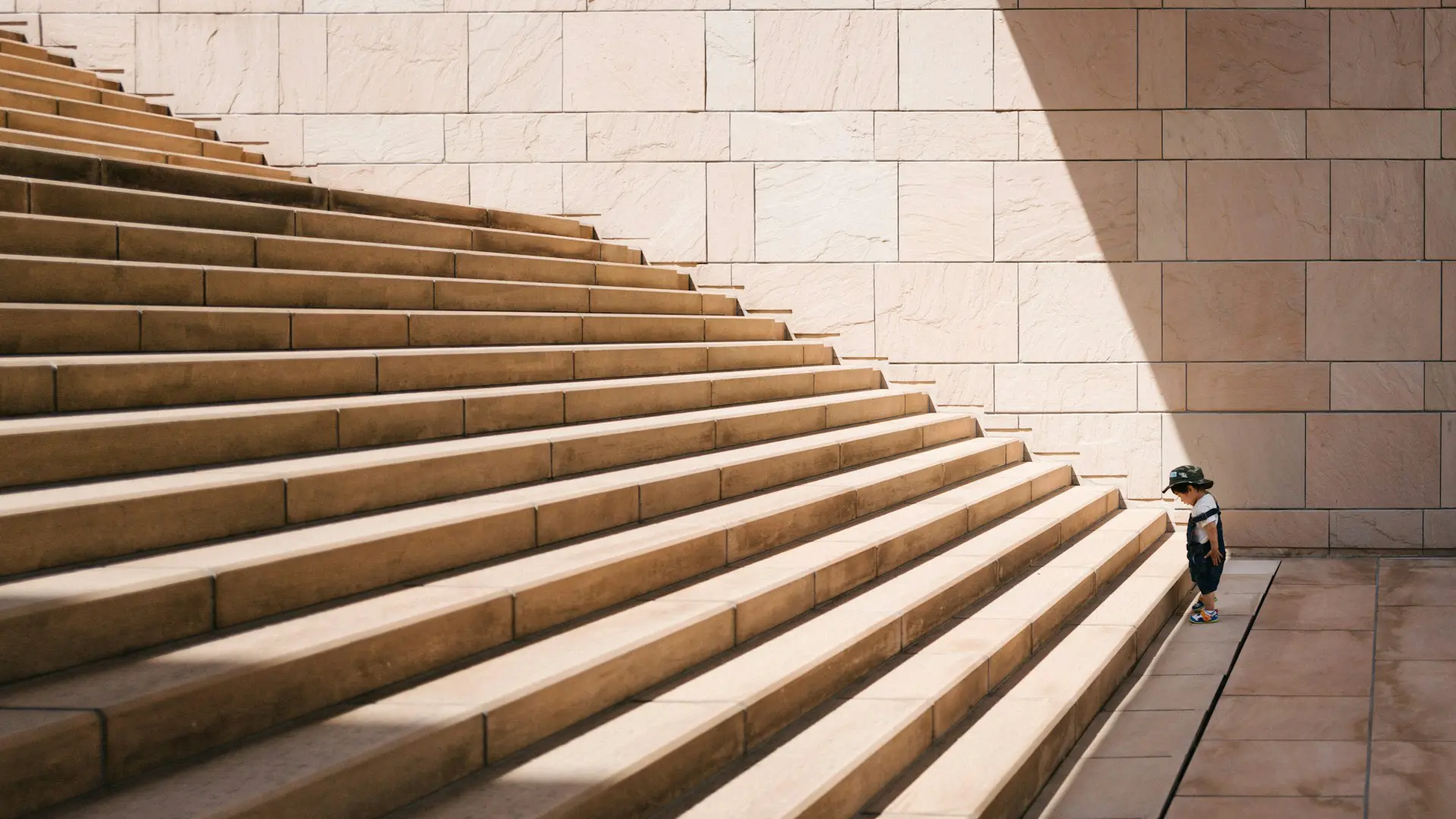 A small child, wearing a hat and dark clothing, stands at the bottom of a large staircase, looking up at the steps. The smooth, light-colored stone background and sunlight create dramatic shadows, capturing not just a journey upward but also different types of stress faced in life.