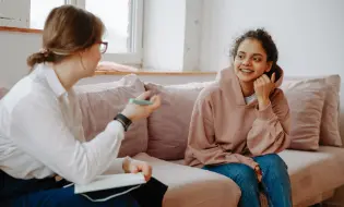 Two people sit on a light-colored sofa, engaged in conversation. One, holding a pen and notebook, listens attentively. The other, wearing a hoodie and jeans, smiles and gestures with her hand. They are in a well-lit room with a window in the background, discussing PTSD treatment options.