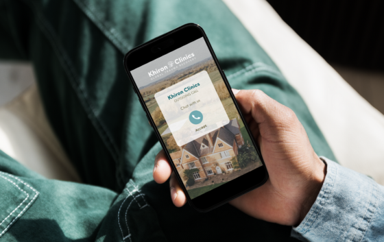 A person wearing green pants and a light denim shirt is sitting and holding a smartphone. The phone screen displays an incoming call from "Khiron Clinics," a renowned trauma clinic, with options for accepting the call. The app’s interface shows a country house in the background.