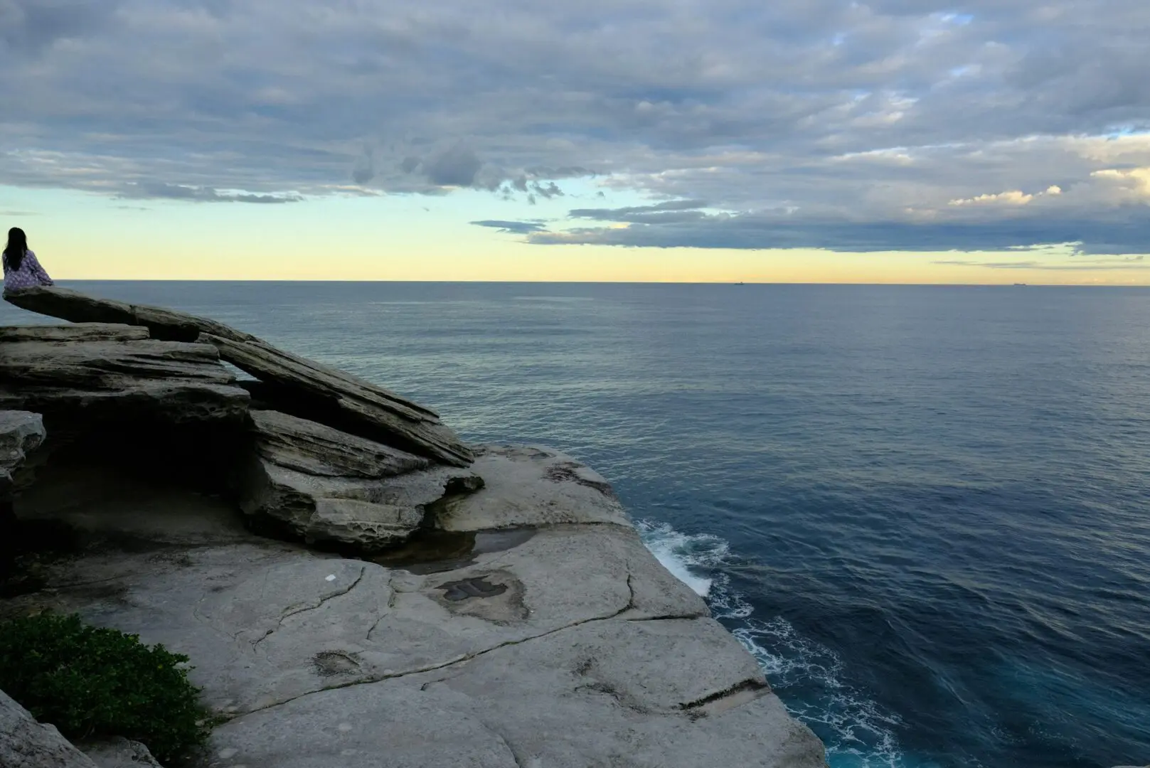 A person sits at the edge of a rocky cliff overlooking a vast ocean, lost in thought. The sky above is partly cloudy with a hint of sunset hues on the horizon. The calm ocean blends seamlessly with the horizon, creating a serene view that starkly contrasts their memories of childhood neglect.