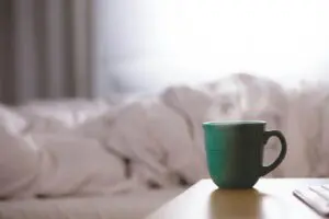 A green mug sits on a wooden table in the foreground, with a blurred, bright bedroom scene featuring unmade white bedding in the background. The cozy morning atmosphere contrasts with the remnants of nightmares lingering from sleep paralysis.