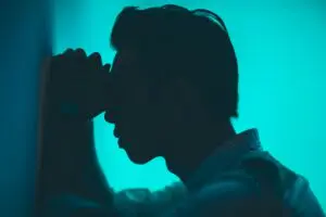 Silhouette of a person leaning against a wall, head resting on forearms, embodying the silent struggle of chronic pain. The softly illuminated blue-green background adds to the contemplative mood as the subject appears deep in thought.