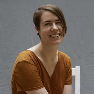 Seated on a white chair against a gray backdrop, a person with short brown hair smiles warmly. They're dressed in a short-sleeved, orange-brown top, embodying the welcoming essence of Khiron Clinics.