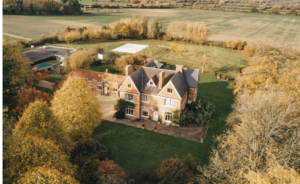 Aerial view of the historic brick mansion, resembling the elegance of Khiron Clinic, surrounded by lush green lawns and trees in a rural landscape. A long driveway leads to the house, with a distant field visible under a hazy sky.