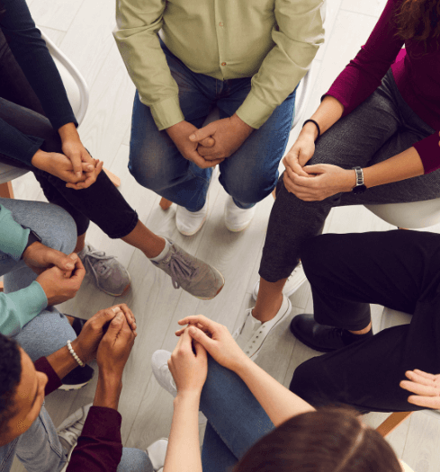 A group of people sitting in a circle on chairs, seen from above, with their hands folded or resting on their laps, suggesting a meeting or support group discussing topics like What is Trauma.