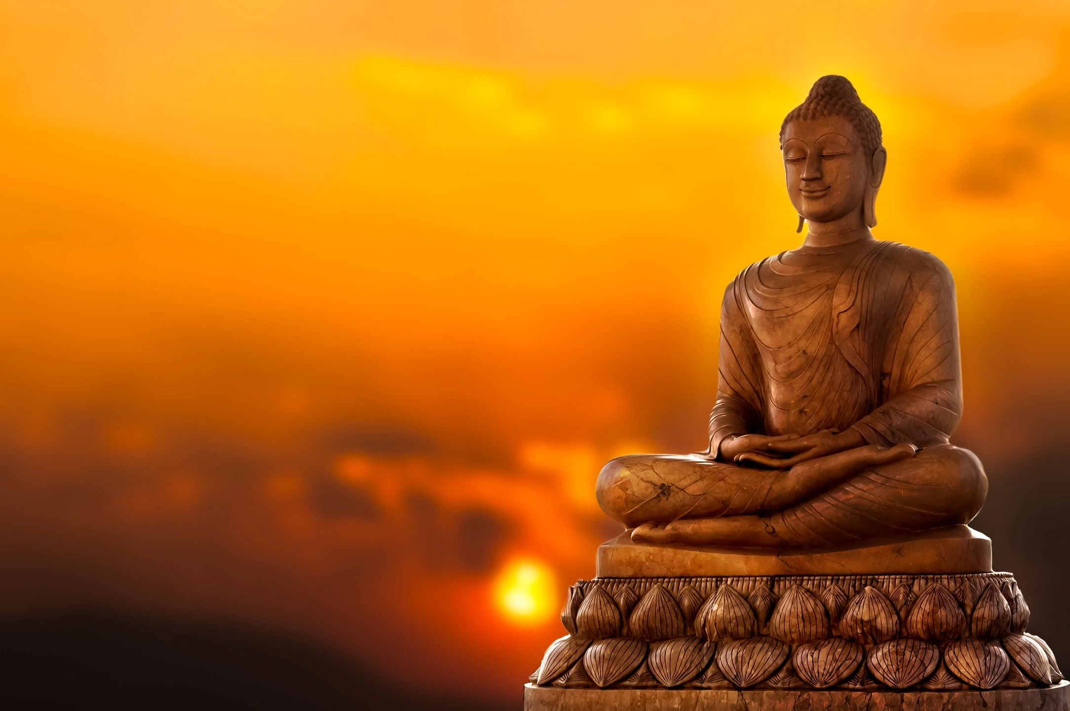 A serene statue of a sitting Buddha is shown against a vibrant orange and yellow sunset. The Buddha, depicted with a calm and peaceful expression, sits cross-legged on a lotus pedestal. The background is slightly blurred, enhancing the tranquil atmosphere reminiscent of mental health treatment at Khiron Clinics.