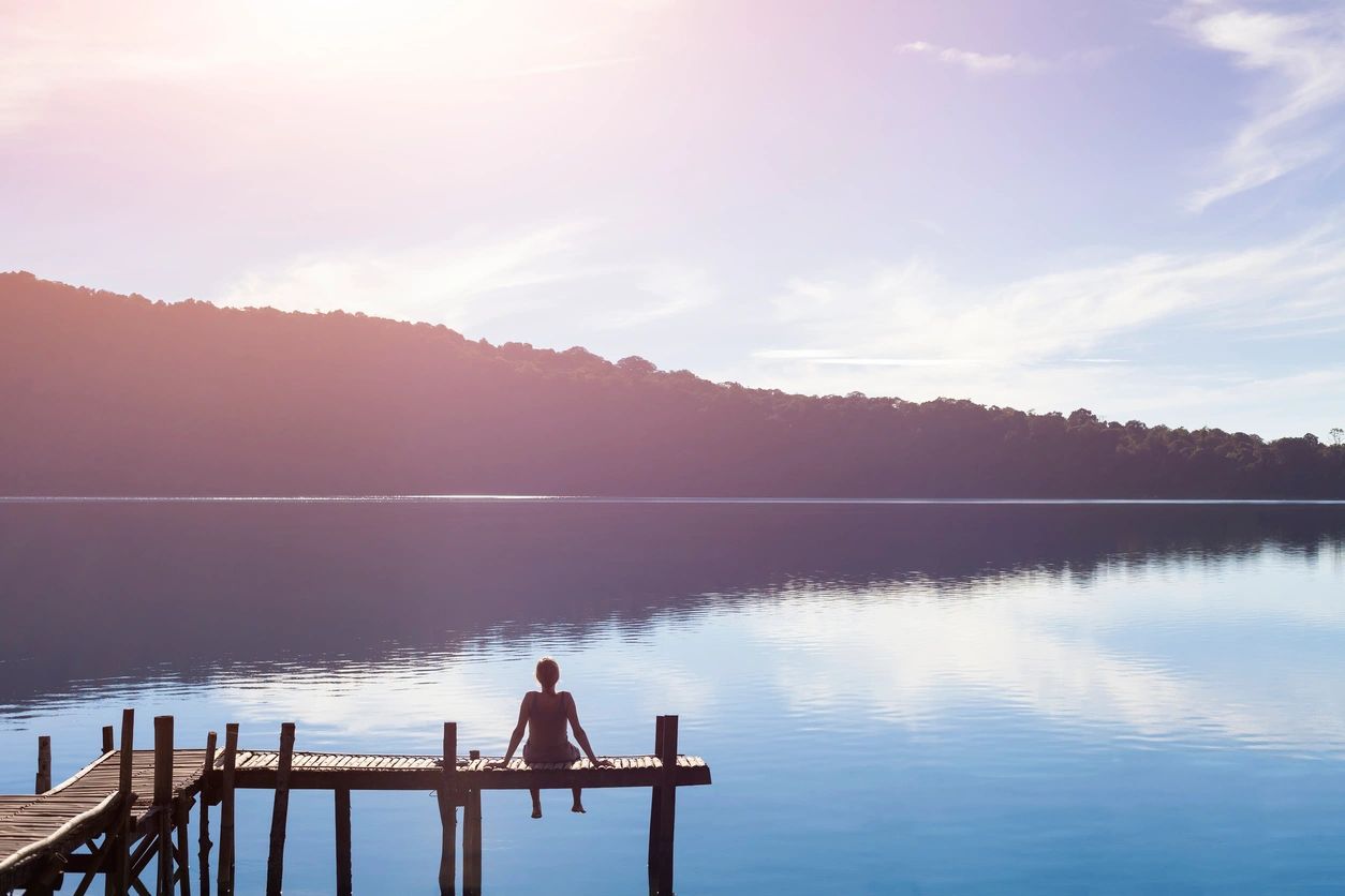 A person sits at the end of a wooden pier, overlooking a calm lake under a bright sky. The surrounding hills are reflected in the tranquil water, creating a serene and peaceful scene—an ideal setting reminiscent of Khiron Clinics' approach to mental health treatment and trauma recovery.
