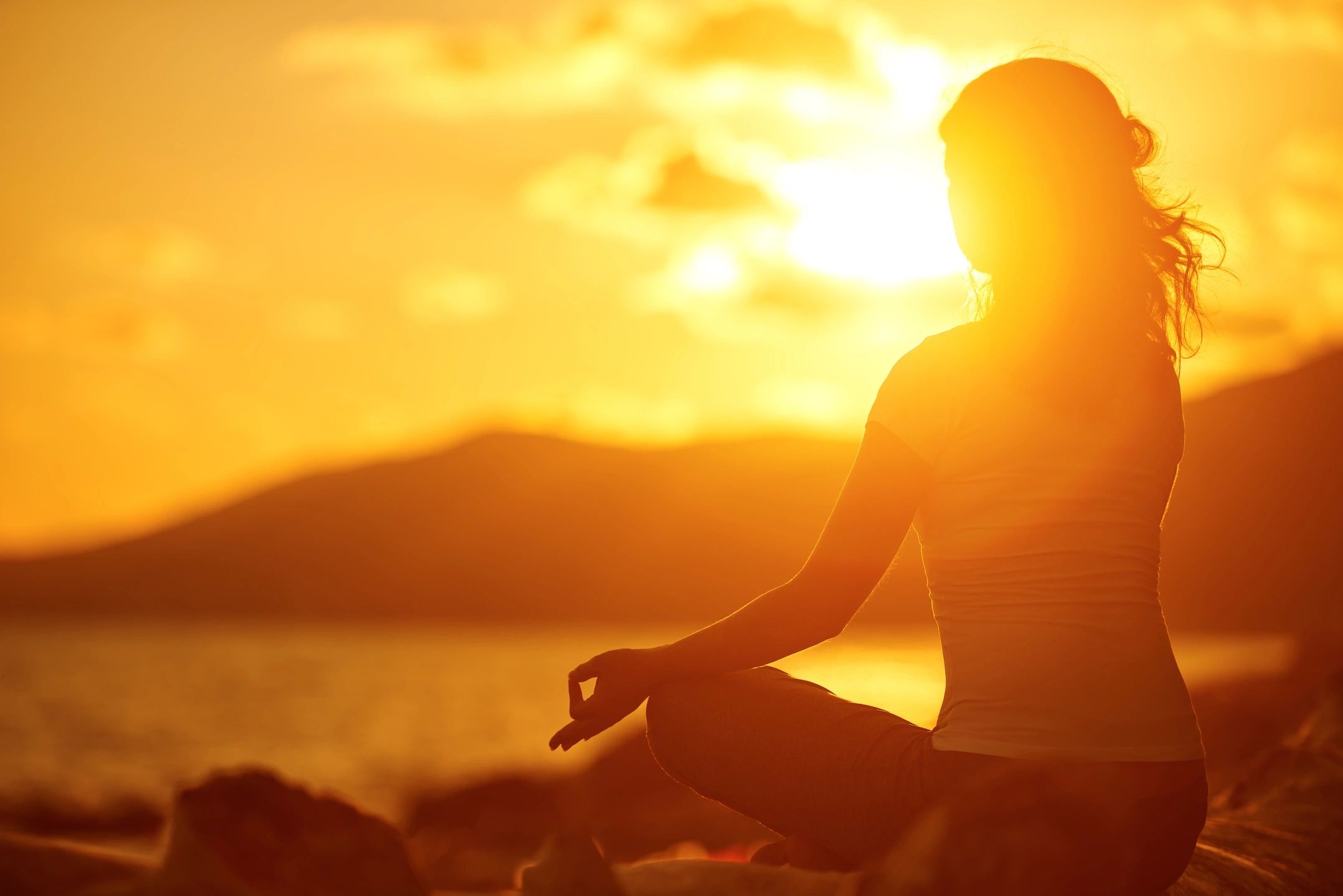 A person sits in a meditative pose by the water during sunset. The bright sunlight creates a glowing silhouette, highlighting their peaceful posture. This tranquil moment serves as a natural mental health treatment against the backdrop of blurred mountains and a serene, golden sky.
