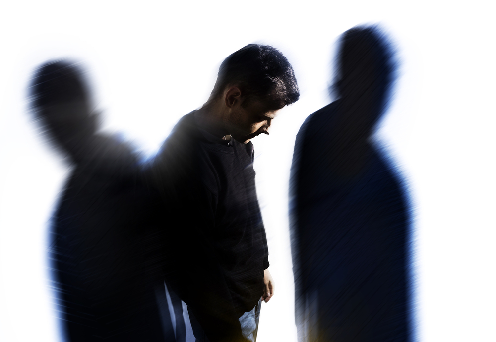 A man in a black shirt stands with his head bowed. Blurred, shadowy figures flank him on both sides against a white background, creating an abstract, almost ghostly appearance. The image conveys a sense of solitude and introspection, reminiscent of the profound journey experienced at Khiron Clinics.