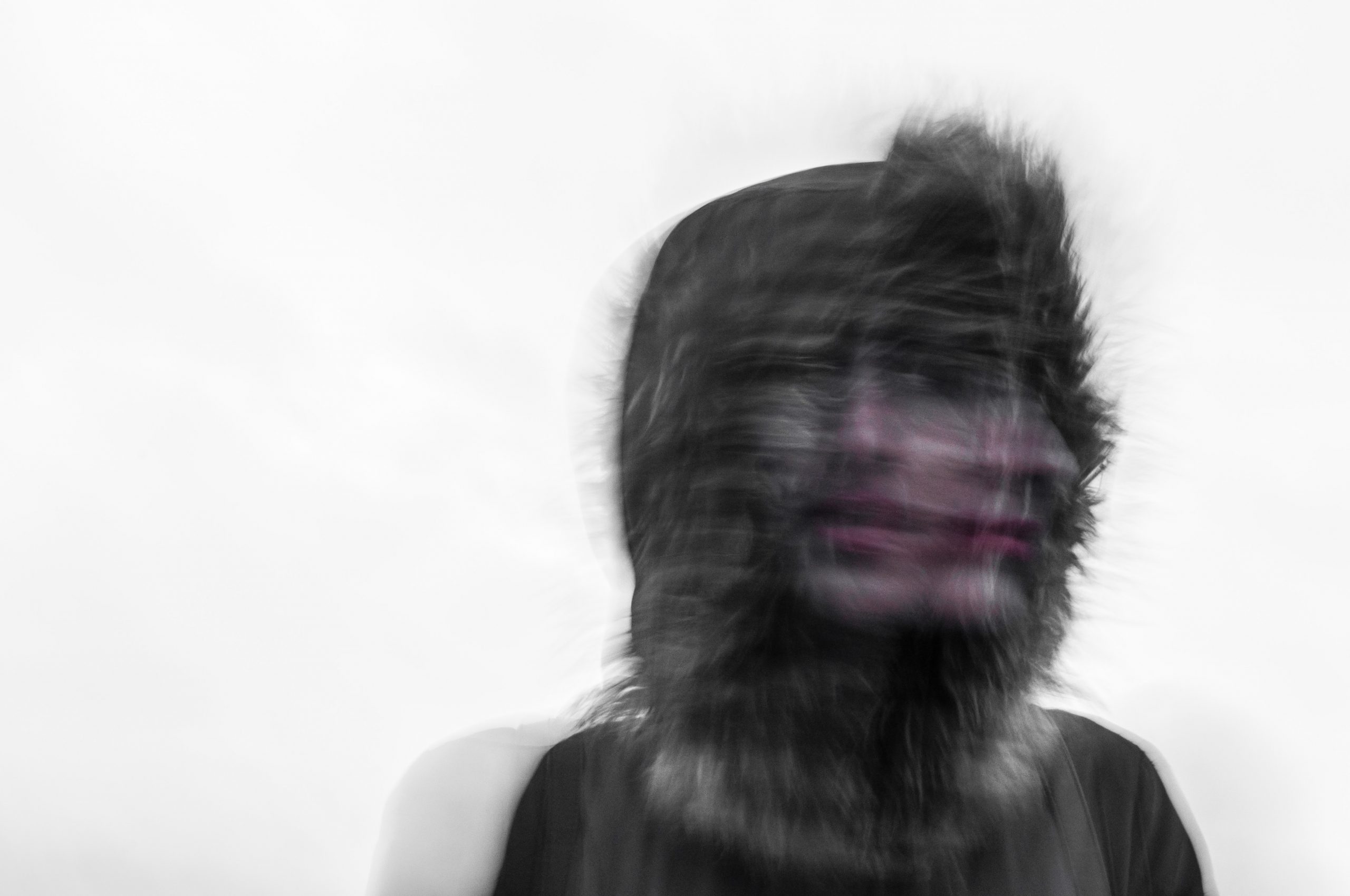 A blurry, black and white image of a person wearing a hooded jacket with fur trim. The person's face appears distorted and doubled, creating a ghostly effect against a plain, light-colored background—evocative of the isolation felt during trauma treatment at Khiron Clinics.