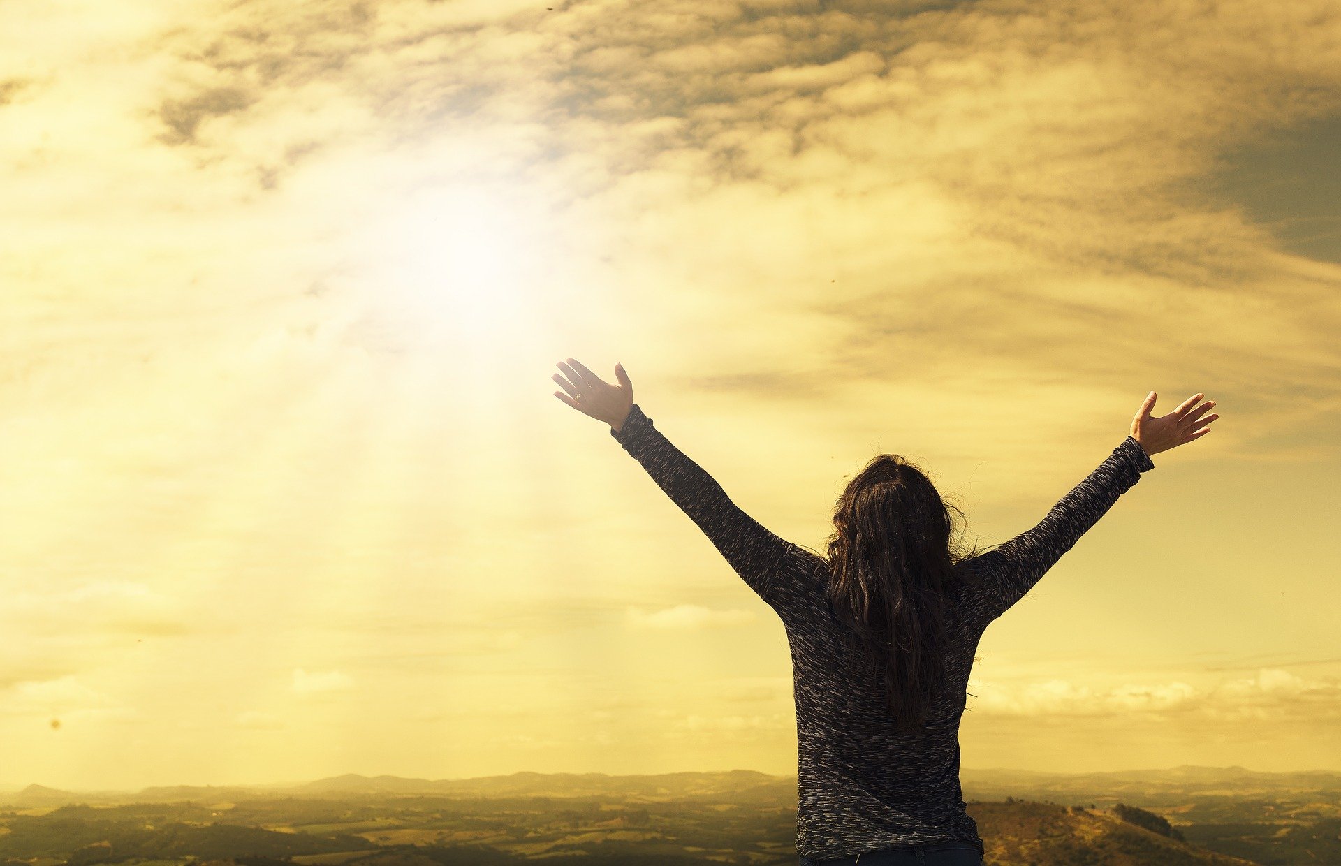 A person with long hair is standing on a hilltop with their arms raised towards the sky. The sun is shining brightly through the clouds, casting a golden hue over the landscape of rolling hills and distant mountains, offering a serene moment of reflection after visiting the trauma clinic.