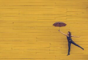 A woman, dressed in black, is playfully holding an open umbrella and appears to be floating against a bright yellow wooden wall. She extends one leg behind her in mid-air, creating a whimsical scene that reminds us how essential moments of joy are even during mental health treatment. The overall tone is cheerful and light-hearted.