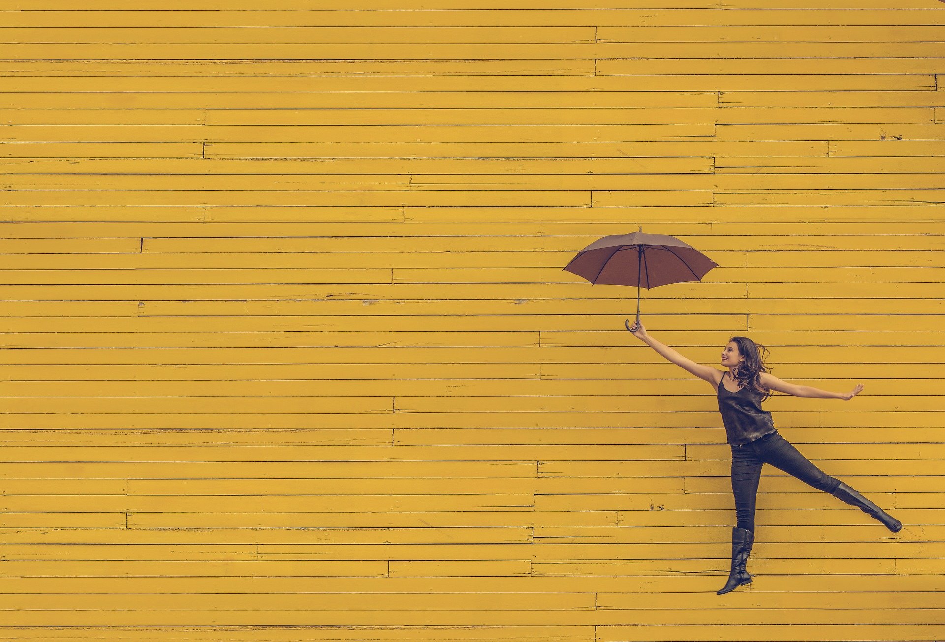 A woman, dressed in black, is playfully holding an open umbrella and appears to be floating against a bright yellow wooden wall. She extends one leg behind her in mid-air, creating a whimsical scene that reminds us how essential moments of joy are even during mental health treatment. The overall tone is cheerful and light-hearted.