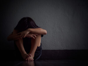 A person with long hair is sitting on the floor, curled up with their arms resting on their knees and head hidden. The scene is dimly lit, creating a somber and reflective mood against a plain, dark background, reminiscent of the introspective moments often experienced in trauma treatment at Khiron Clinics.