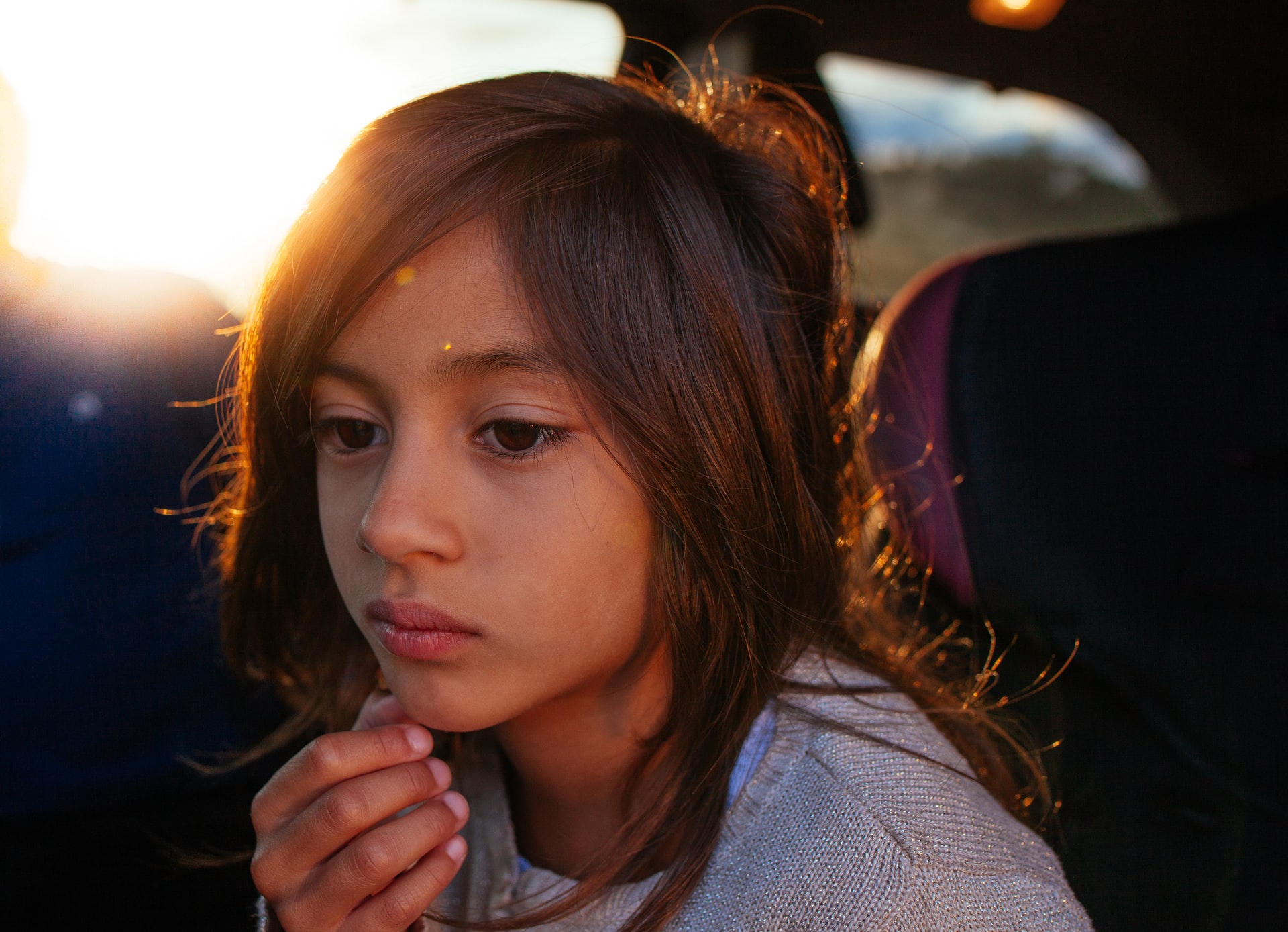 A child with long hair is sitting in a car seat, gazing thoughtfully out of the window. Warm sunlight illuminates the child's face from behind, casting a gentle glow and creating a serene atmosphere. The child's hand rests lightly against their chin, perhaps reflecting on experiences from Khiron Clinics.