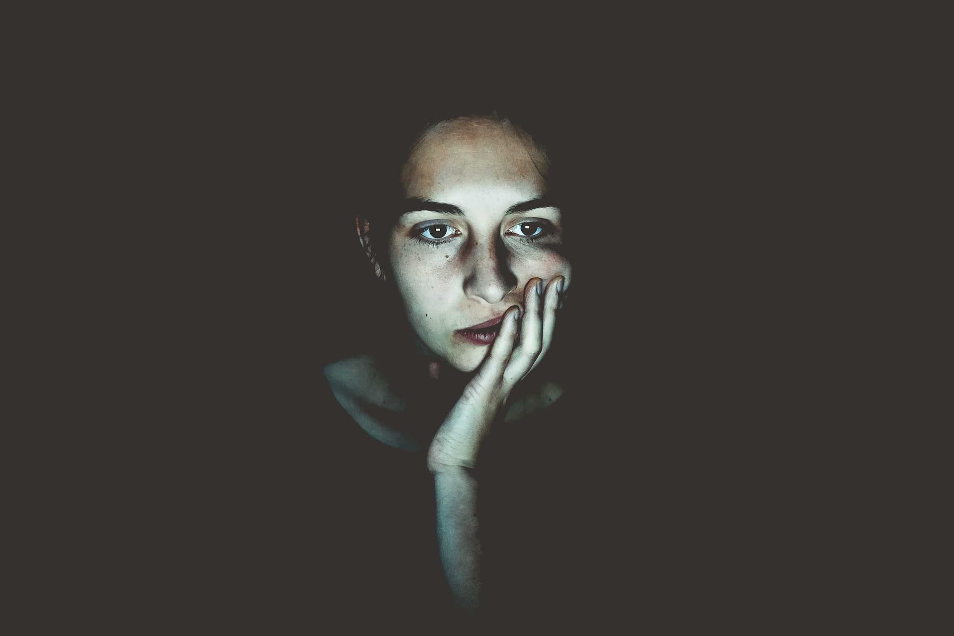 A person with a contemplative expression holds their hand to their face. The low, dim lighting highlights their face against a dark background, creating a dramatic effect. The person's eyes appear focused, suggesting they might be reflecting on trauma treatment, adding to the pensive mood of the image.