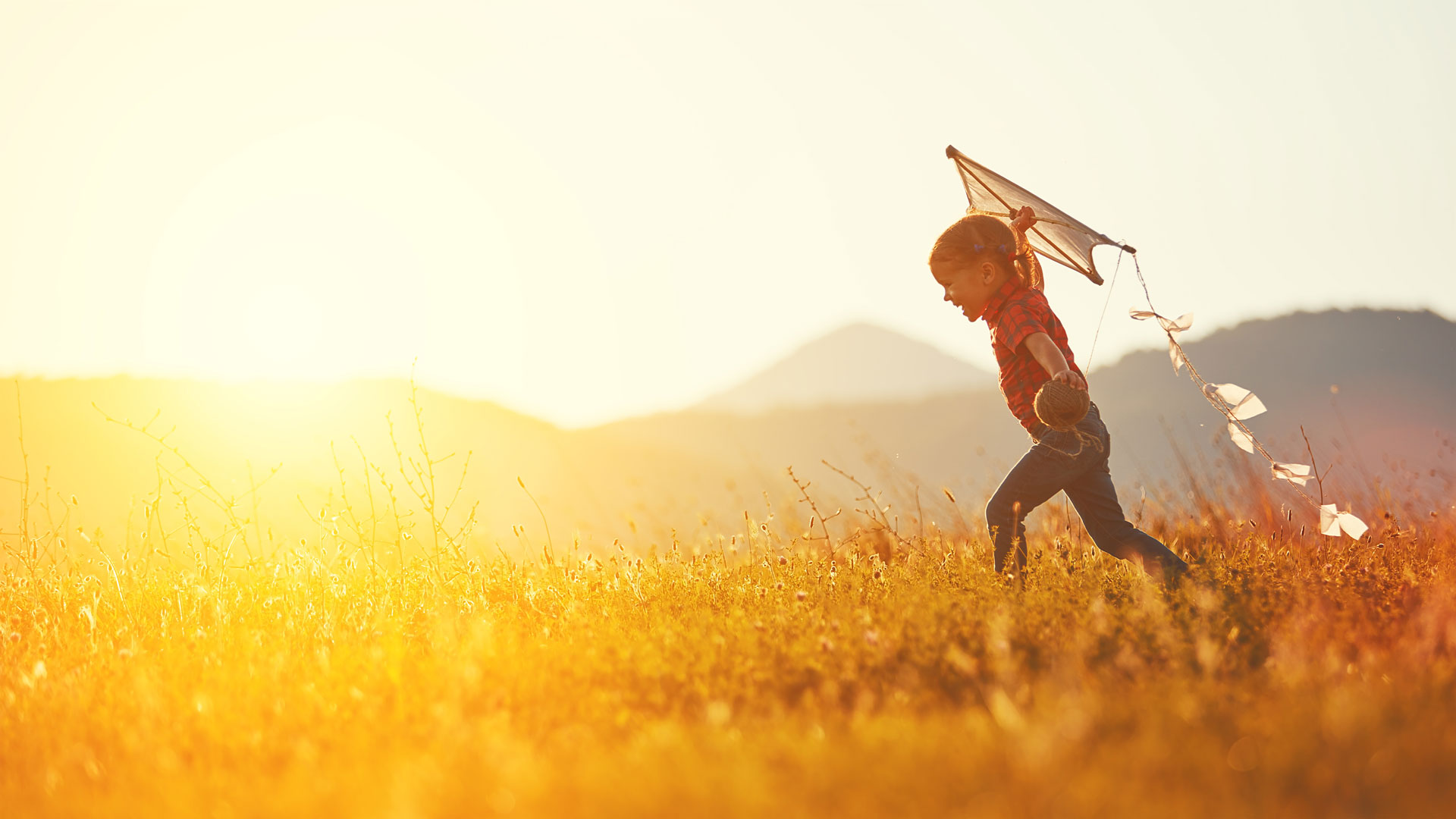 A child runs through a sunlit field with a kite trailing behind. The distant mountains and warm, golden glow of the setting sun create a serene, joyful scene. The carefree spirit of the moment contrasts sharply with memories often addressed in trauma clinics.