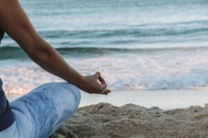 A person sits cross-legged on a sandy beach, facing the ocean. Their hand rests on their knee in a meditation pose, with their thumb and index finger touching. The beach and waves are calm, creating a serene atmosphere ideal for mental health treatment like those offered at Khiron Clinics.