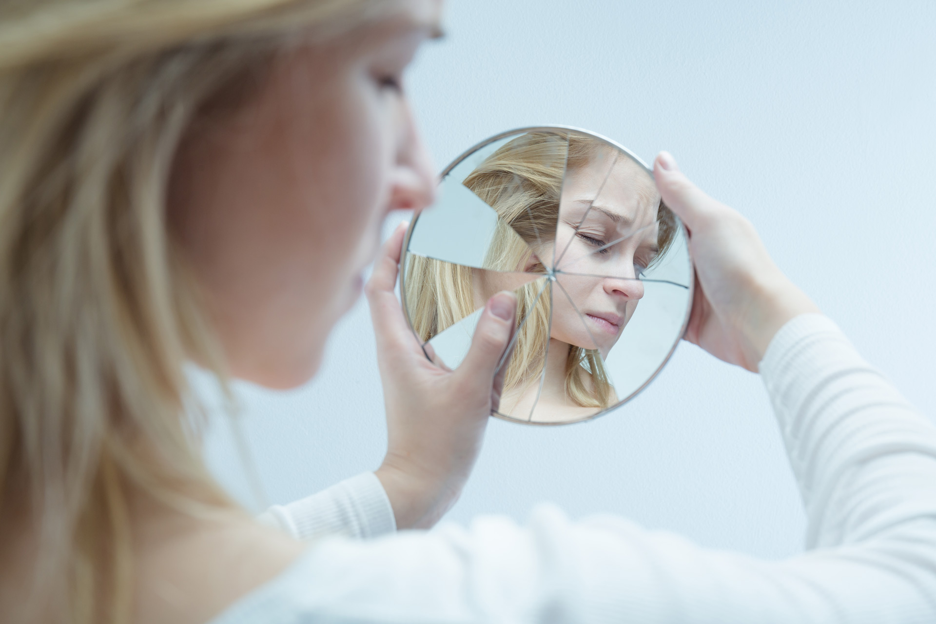 A woman with blonde hair holds a broken circular mirror close to her face, reflecting fragmented pieces of her somber expression. She gazes reflectively at the mirror, as if standing in a trauma clinic like Khiron Clinics, set against a plain, light-colored background.