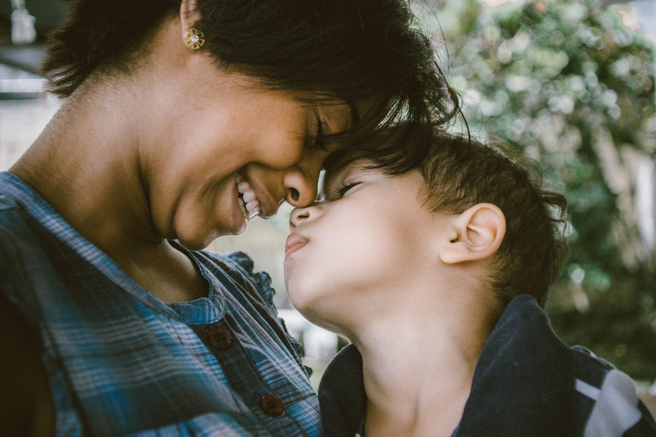 A close-up of a woman and a young boy touching foreheads and smiling. She is wearing a blue plaid shirt, and he has short dark hair and is wearing a navy blazer. They are outdoors with greenery in the background, sharing a tender moment after visiting the trauma clinic for PTSD treatment.