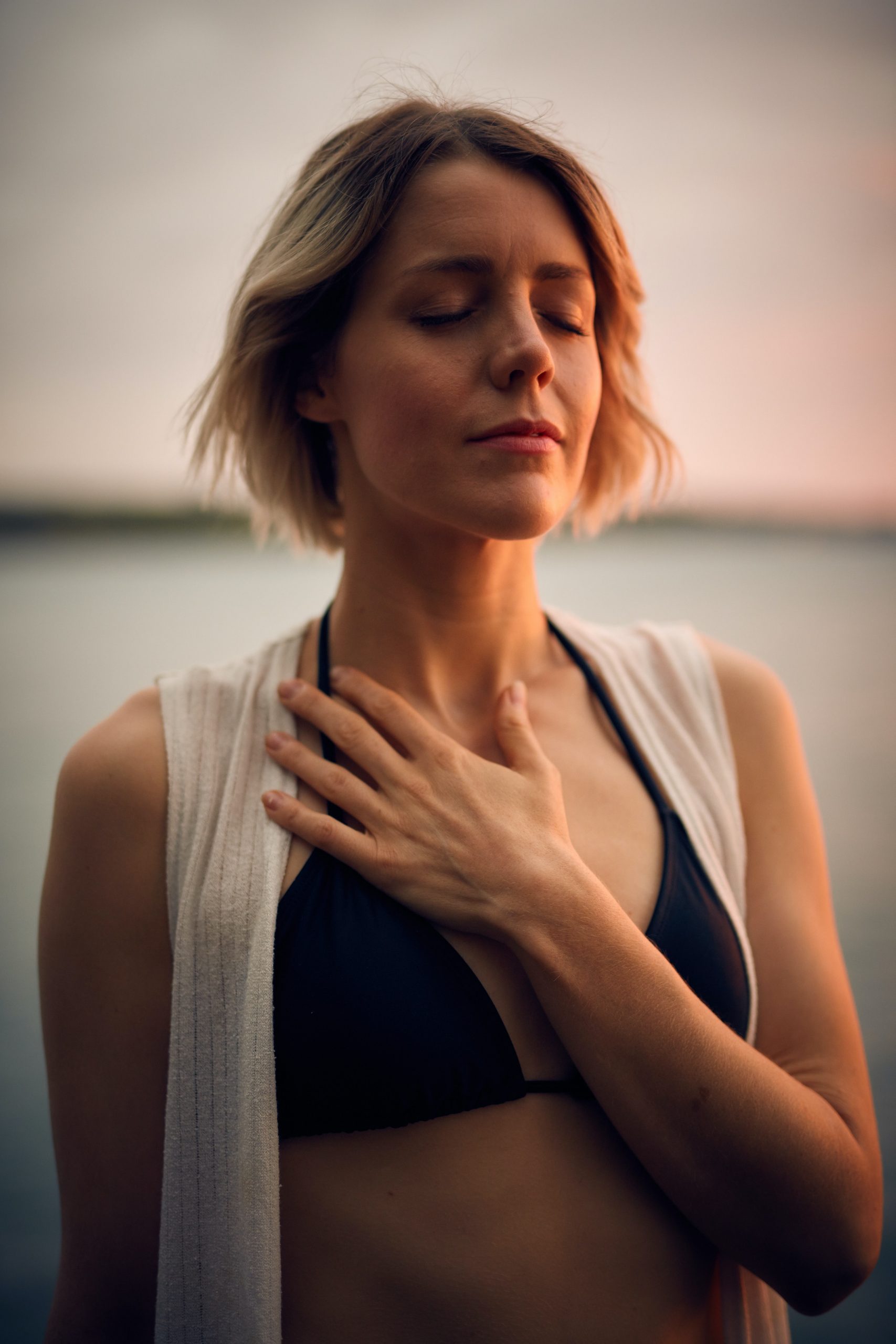 A woman stands outdoors by the water, with her eyes closed and one hand placed on her chest. She has short blonde hair and is wearing a black bikini top and an open white cover-up. The serene backdrop of the sunset over the tranquil water underscores her peaceful moment, reminiscent of trauma treatment at Khiron Clinics.