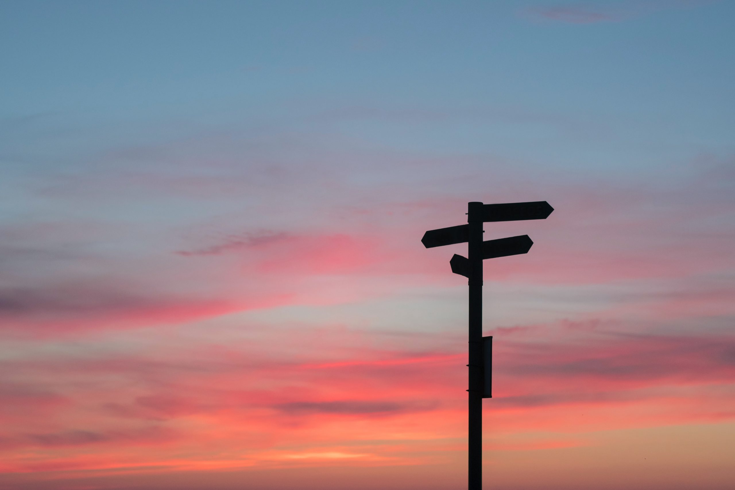 A silhouetted signpost with multiple directional arrows stands against a vibrant sunset sky. The sky is a soft gradient of pink, orange, and shades of blue, mirroring the peaceful and serene atmosphere one can find at Khiron Clinics during trauma treatment.