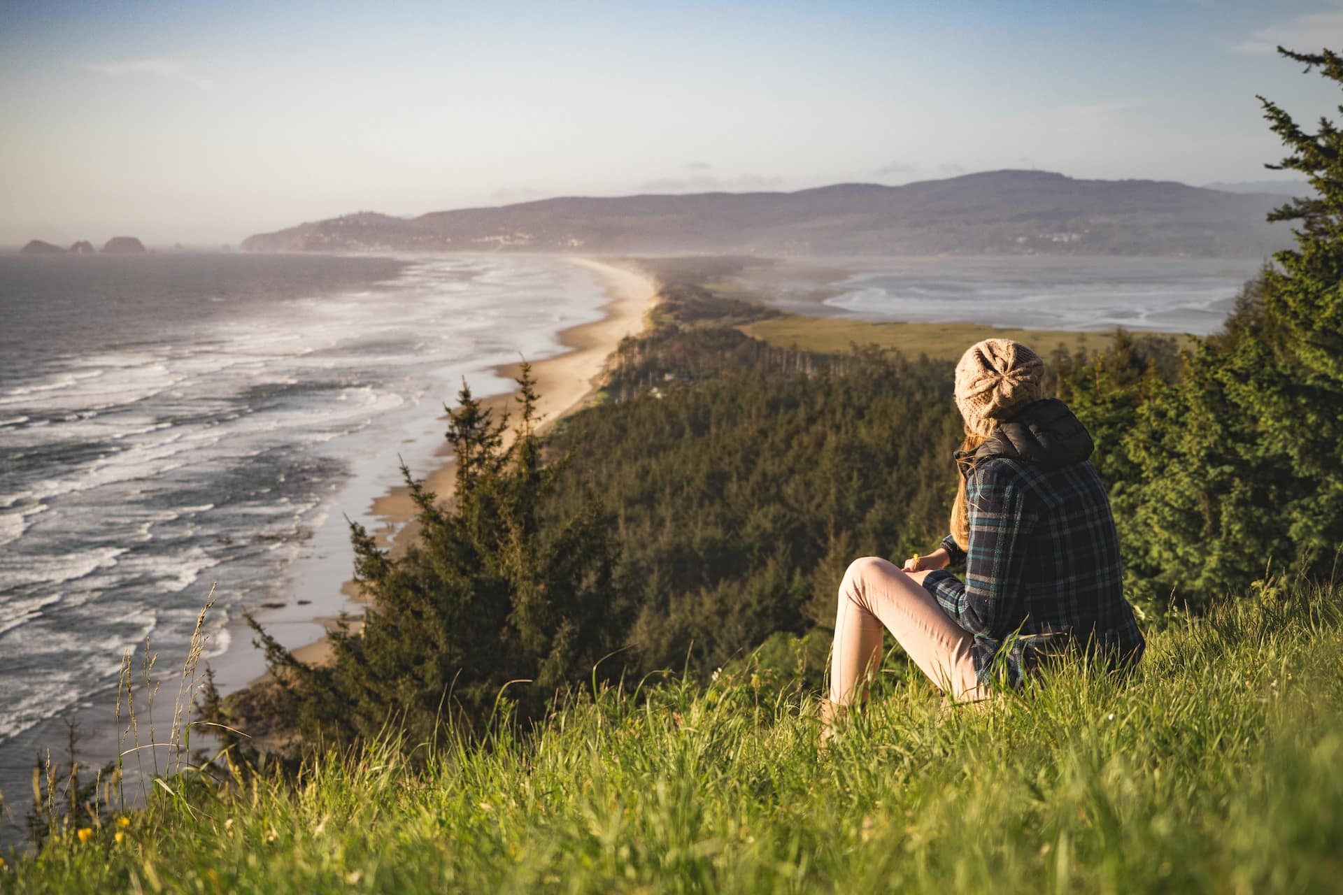 A person wearing a beanie and checkered shirt sits on a grassy hill, overlooking a scenic coastline with waves crashing onto a long stretch of beach. The landscape, perfect for mental health treatment, is filled with dense forests and distant mountains under a clear sky.