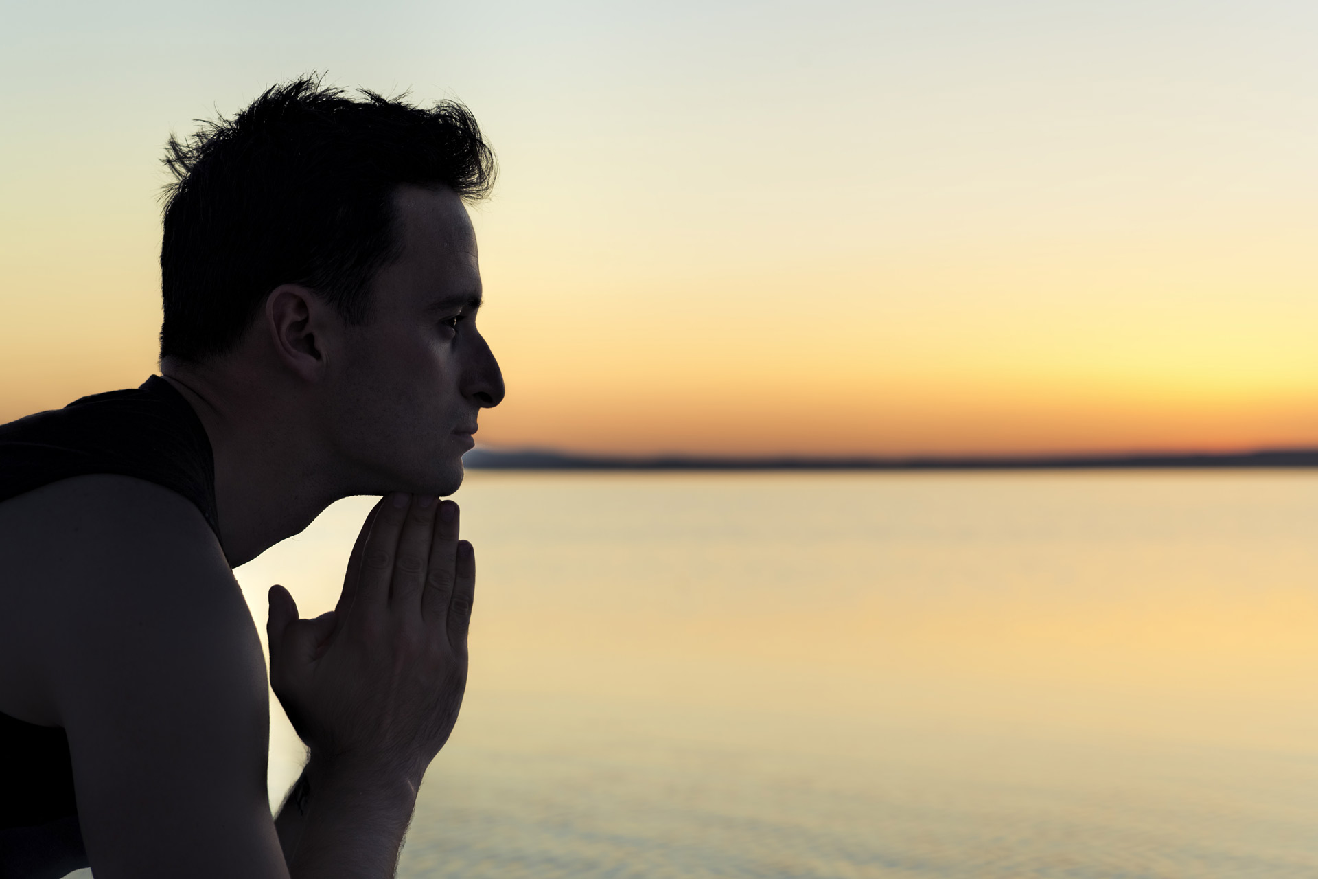 A person, seen in profile, holds their chin in their hands while looking pensively at a serene sunset over a calm body of water. The sky is a gradient of soft yellows and oranges transitioning into dusk, reflecting the healing environment provided by Khiron Clinics.