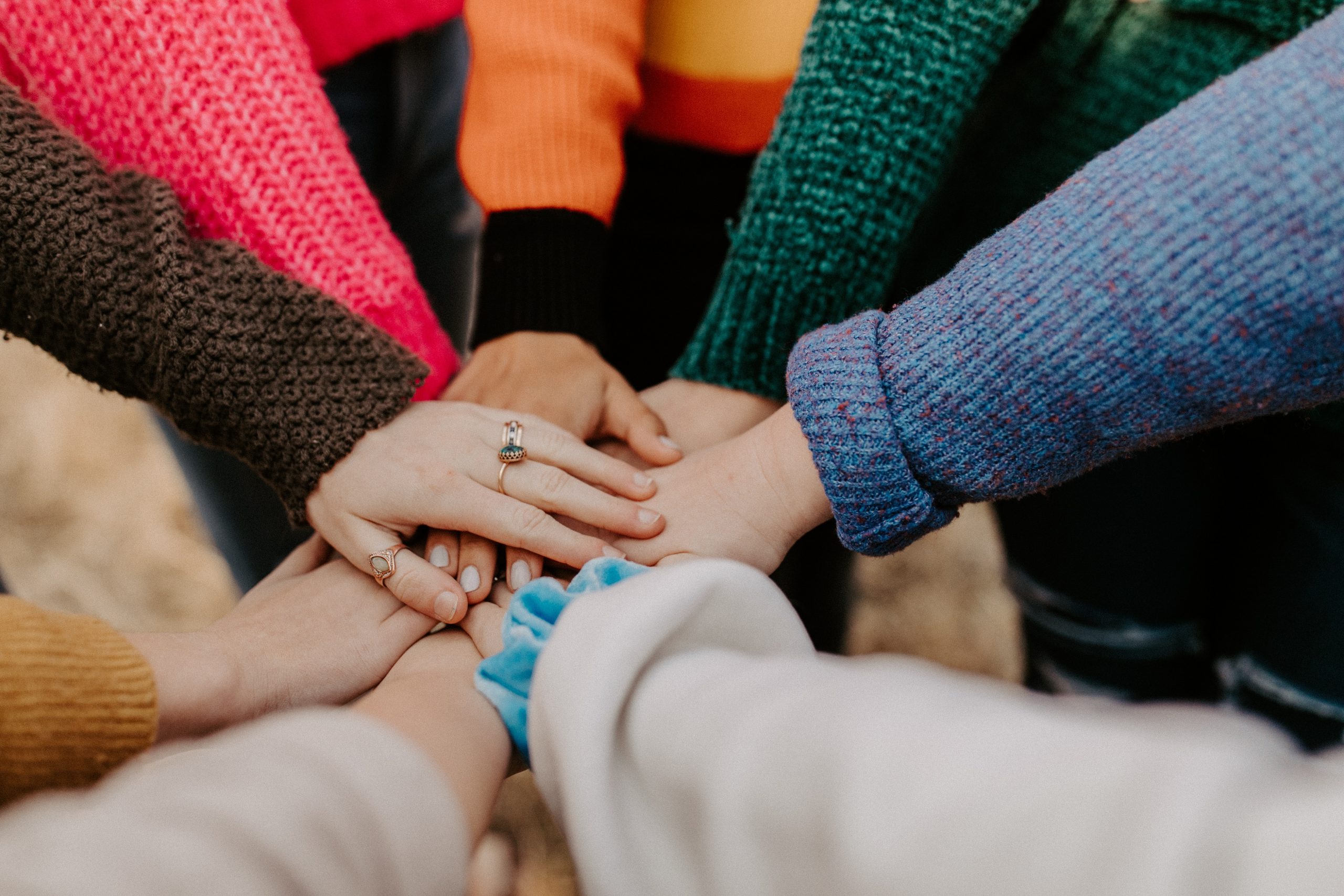 A group of people, each wearing different colorful sweaters, place their hands together in the center of the image, symbolizing unity and teamwork. With the background blurred to highlight the vibrant sweaters and hands, this scene captures the spirit of community at Khiron Clinics where mental health treatment is our priority.